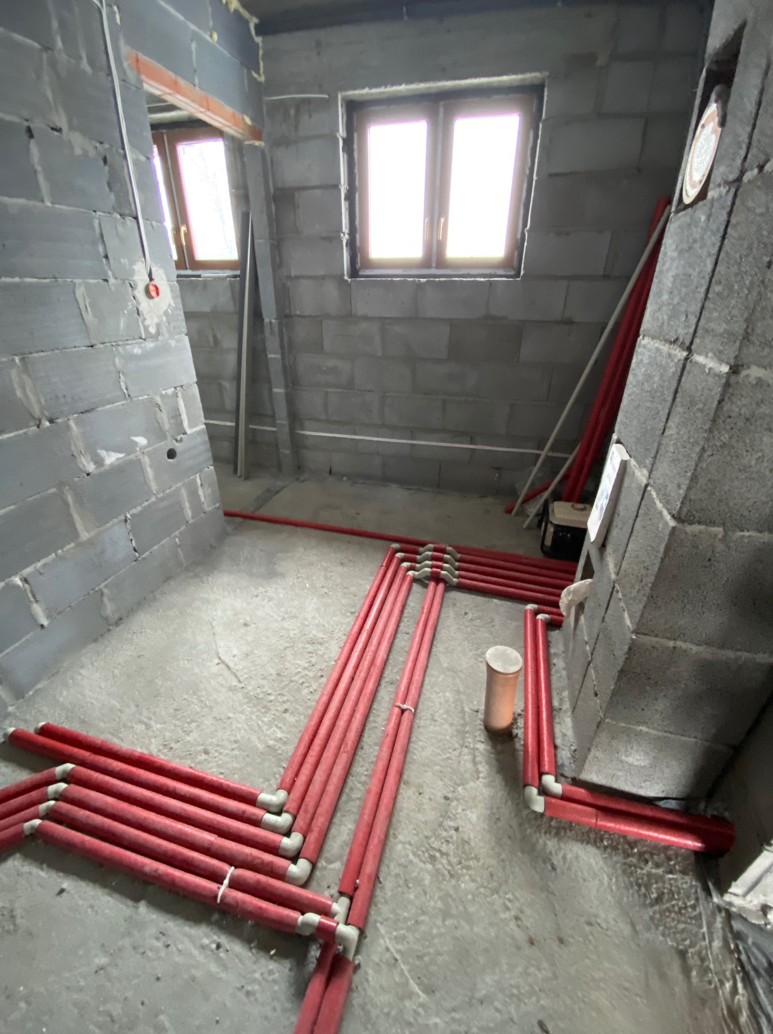 Surowy stan: czerwone rury instalacji grzewczej ułożone na szarej, betonowej podłodze w pomieszczeniu z bloków betonowych, widoczne okna i fragmenty ścian.