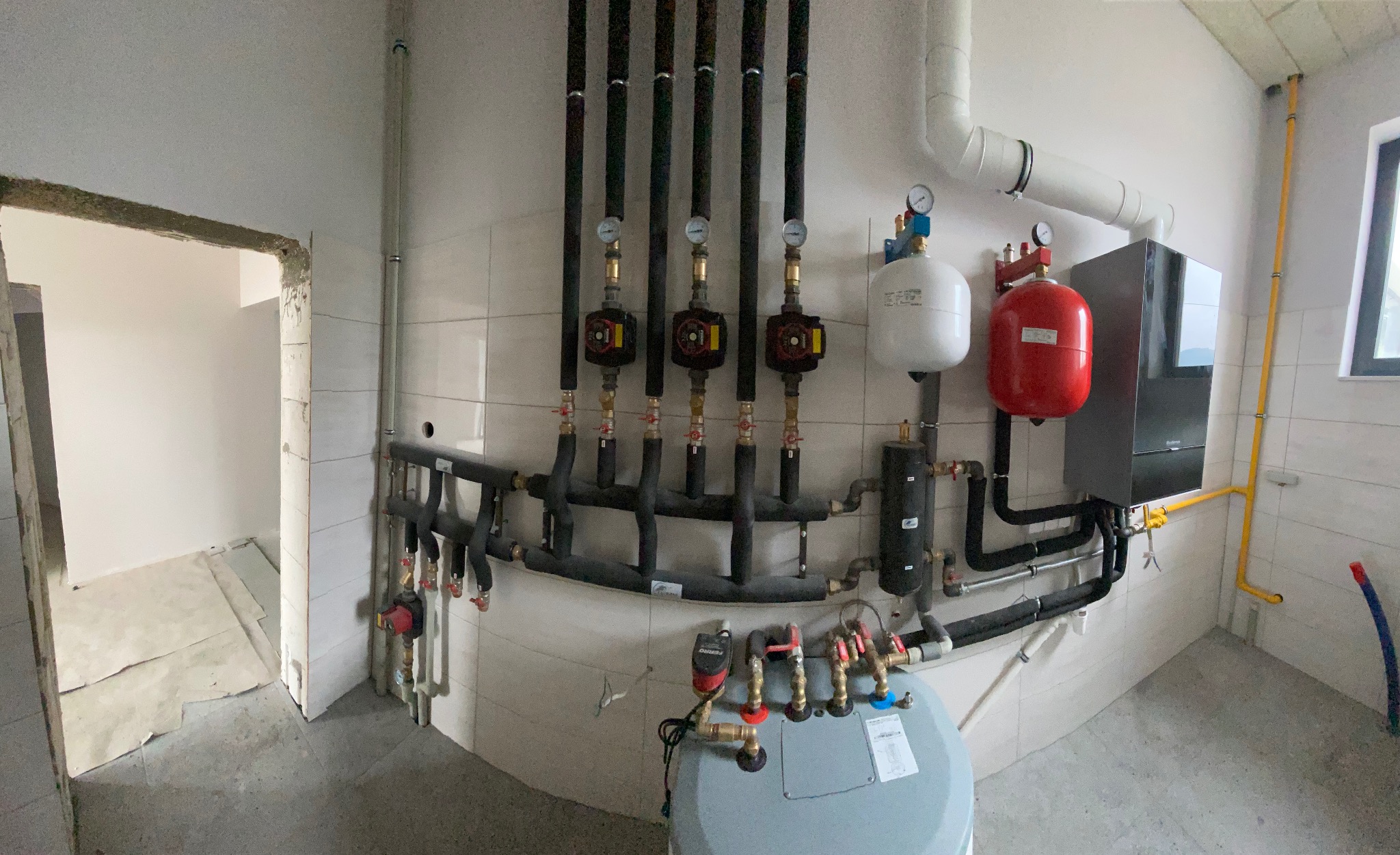 Nowoczesna kotłownia w trakcie montażu: czarny piec Buderus, czerwony zbiornik, biały ekspansomat, rozdzielacze z pompami obiegowymi na ścianie z białymi płytkami, żółta instalacja gazowa, widok...