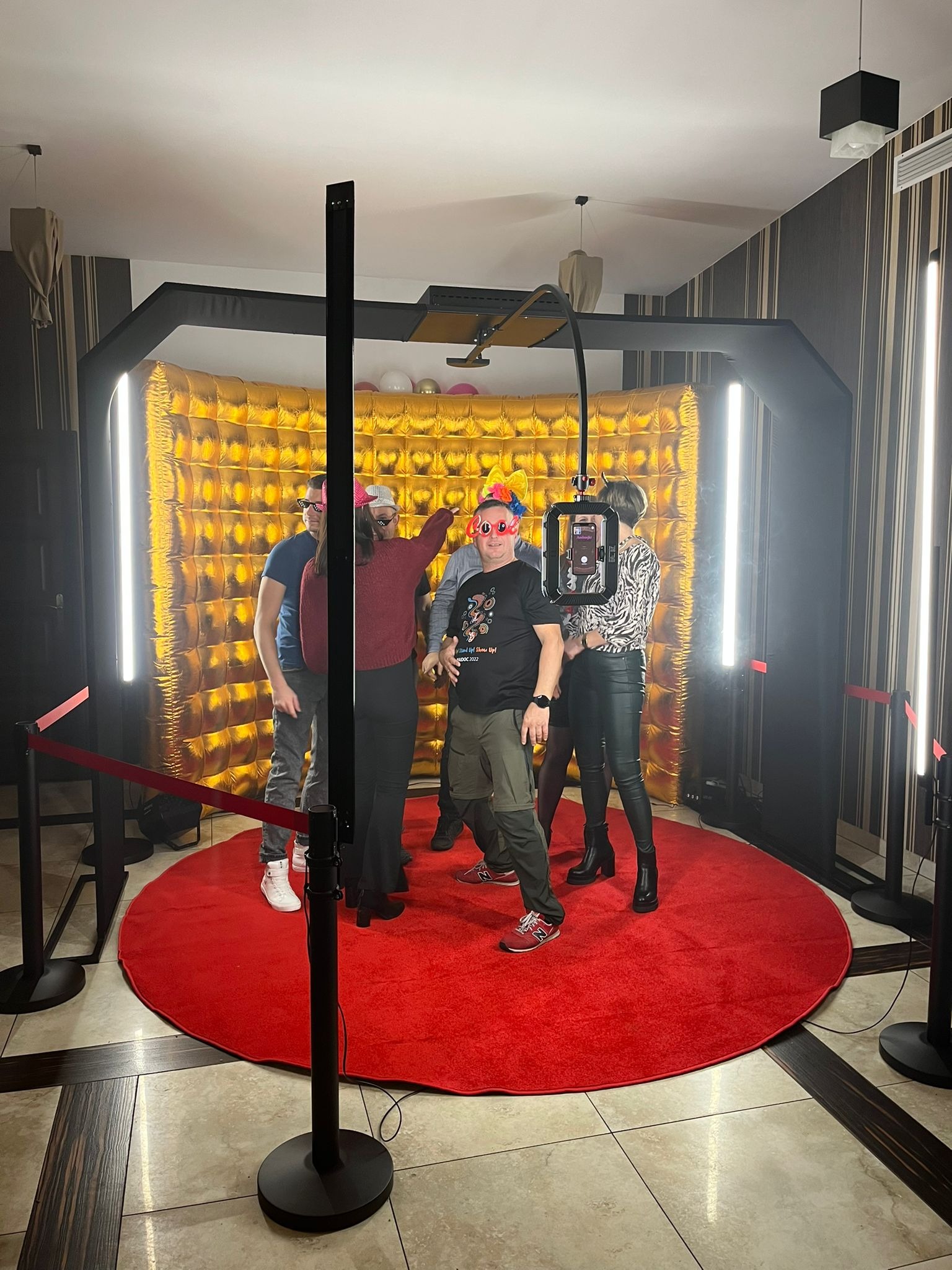 Grupa osób pozuje w fotobudce z złotym, błyszczącym tłem i czerwonym dywanem. Jeden z mężczyzn ma na sobie zabawne, czerwone okulary w kształcie serc.