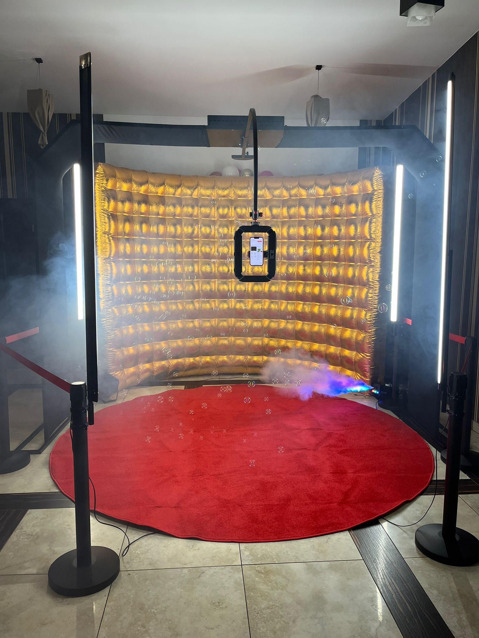 Instalacja z złotym, pikowanym tłem, czerwonym dywanem, oświetleniem LED i wytwornicą baniek mydlanych, z telefonem zamontowanym na statywie, tworząca scenerię do zdjęć.