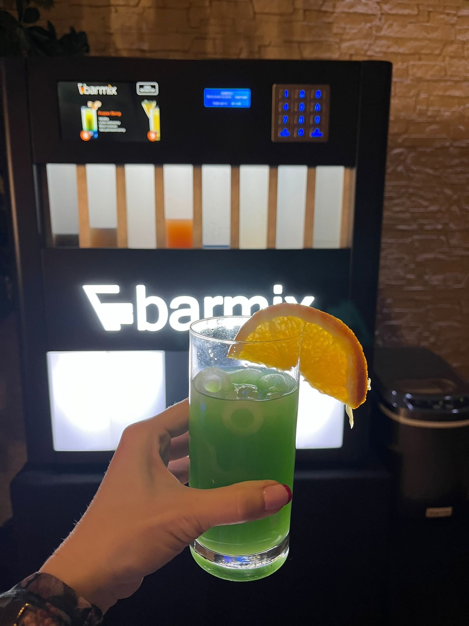 Dłoń trzyma szklankę z zielonym drinkiem i plasterkiem pomarańczy na tle automatu do drinków Barmix.