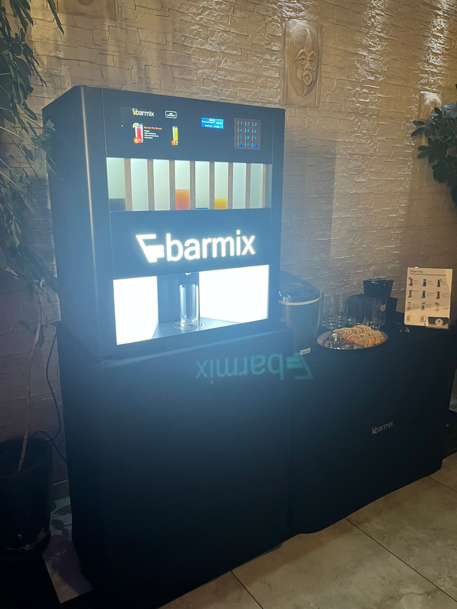Automatyczny bar z logo Barmix, prezentujący menu drinków na ekranie, z pustą szklanką gotową do napełnienia, obok taca z plastrami pomarańczy i ulotka z ofertą drinków.