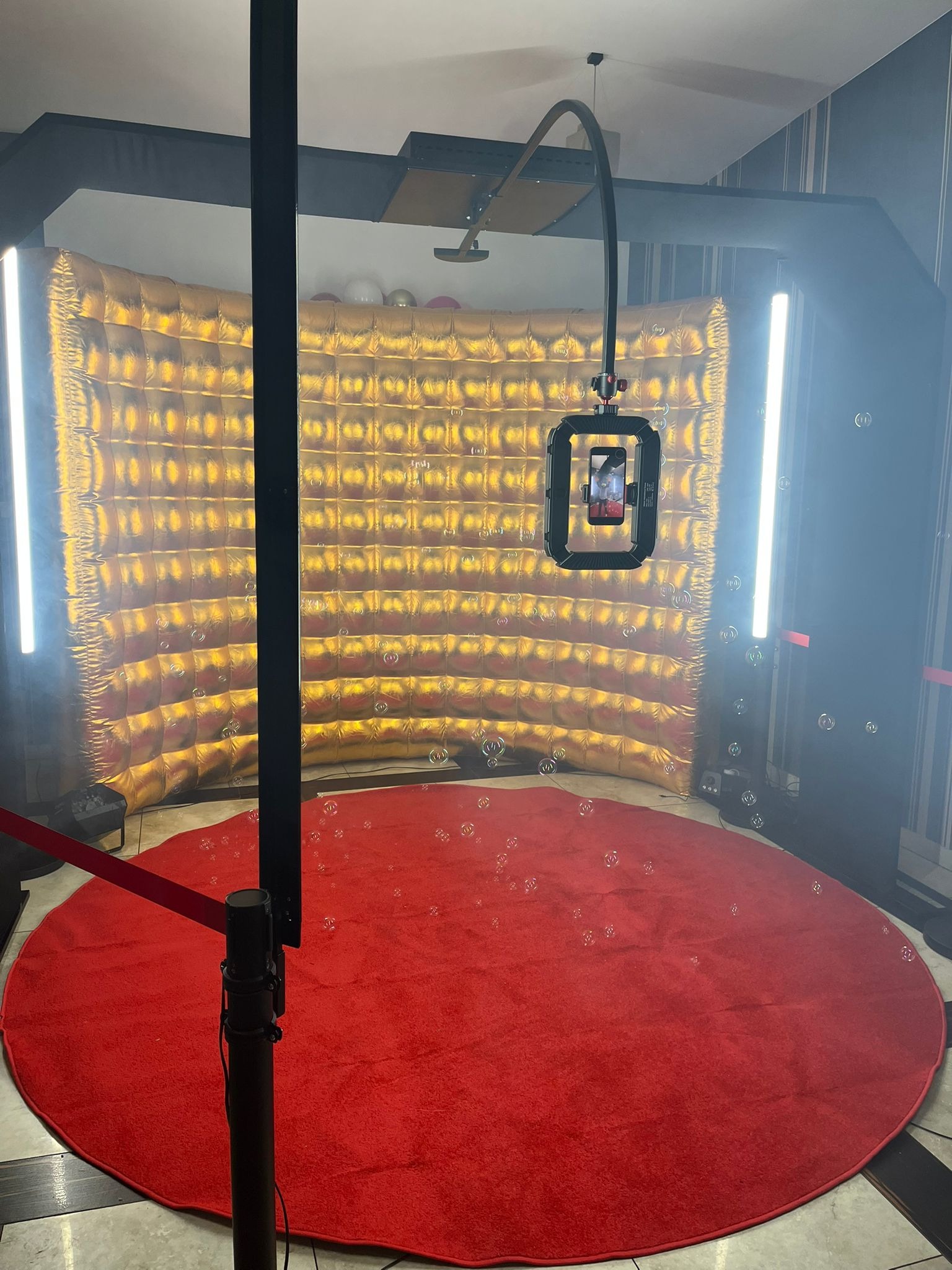 Złota, dmuchana ścianka tła z oświetleniem LED i czerwonym dywanem w przestrzeni do robienia zdjęć, z widocznymi bąbelkami unoszącymi się w powietrzu.