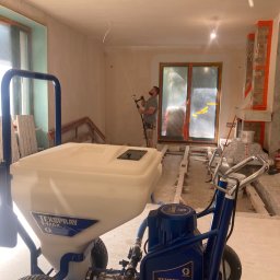 Kamil Błaszczak- Usługi Remontowo-Budowlane - Wnętrze w trakcie prac wykończeniowych, widoczny agregat malarski Texspray T-MAX 506 i pracownik natryskujący ścianę, okno zabezpieczone folią.