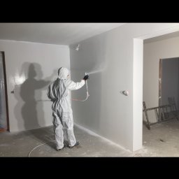 Kamil Błaszczak- Usługi Remontowo-Budowlane - Pracownik w kombinezonie maluje ścianę natryskowo na szaro, widoczny wąż i pistolet natryskowy, w tle surowy stan wykończenia.