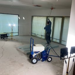 Kamil Błaszczak- Usługi Remontowo-Budowlane - Pracownik w trakcie natryskowego nakładania gładzi na sufit w pomieszczeniu z dużymi oknami zabezpieczonymi folią, widoczny agregat do natrysku i oświetlenie budowlane.