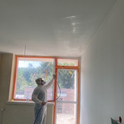 Kamil Błaszczak- Usługi Remontowo-Budowlane - Pracownik w kombinezonie ochronnym maluje natryskowo sufit w pomieszczeniu z oknem zabezpieczonym folią malarską.