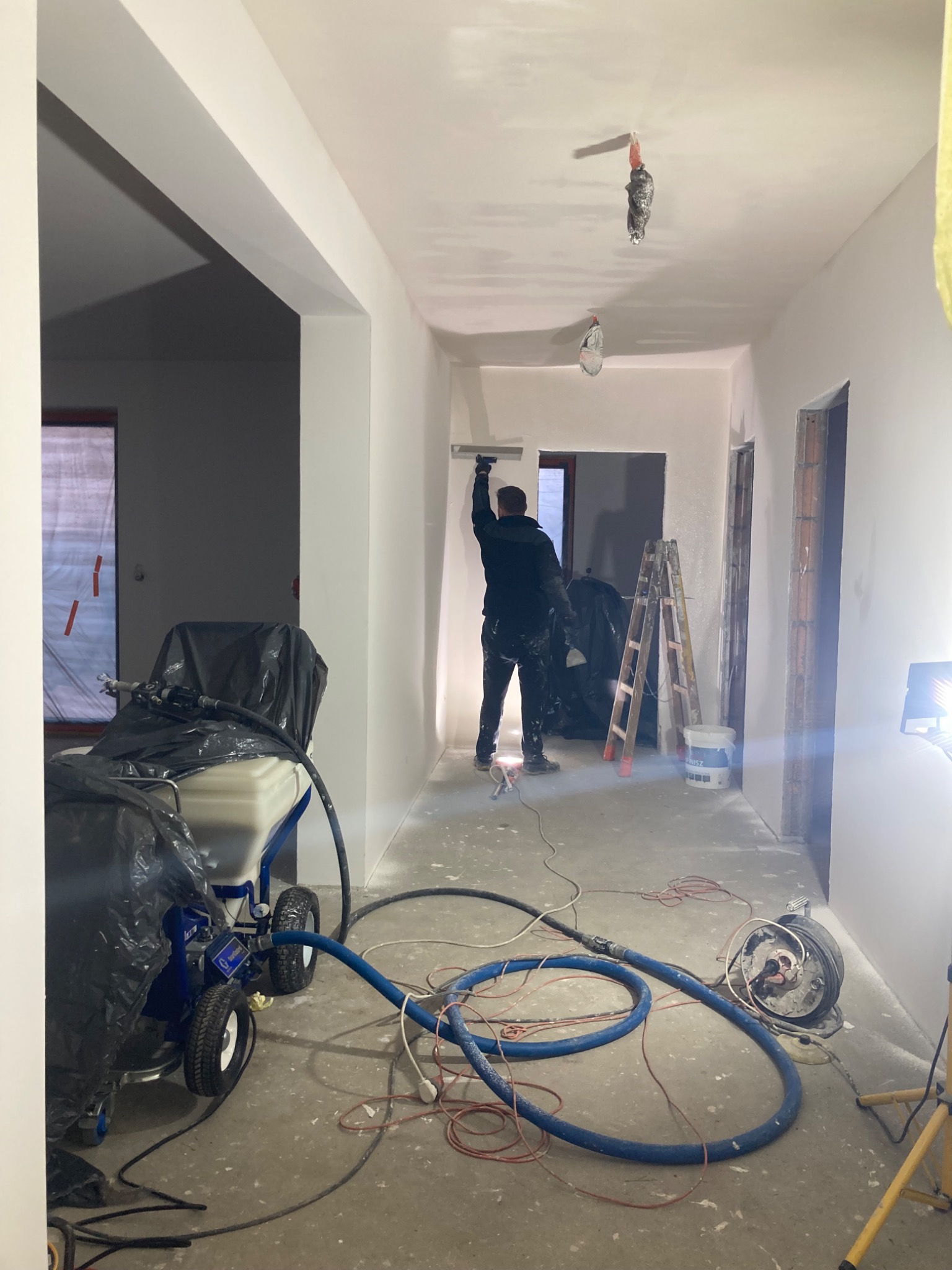 Mężczyzna w ciemnej odzieży szpachluje sufit w długim korytarzu, widoczny agregat szpachlarski przykryty folią, drabina, kable i lampa halogenowa.