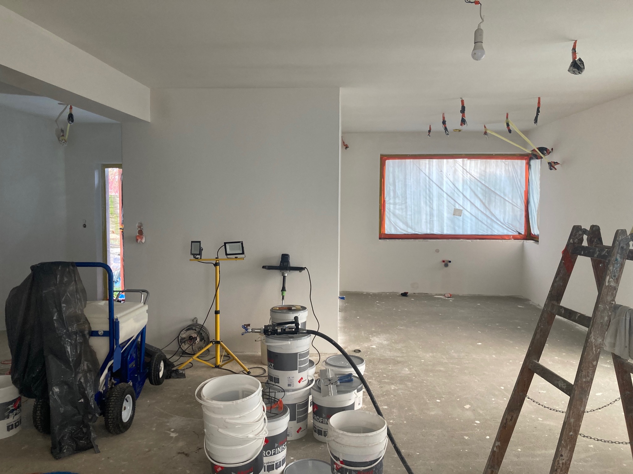 Wnętrze w trakcie remontu: świeżo wyszpachlowane ściany, okno zabezpieczone folią, drabina oparta o ścianę, sprzęt do szpachlowania i wiaderka z farbą.