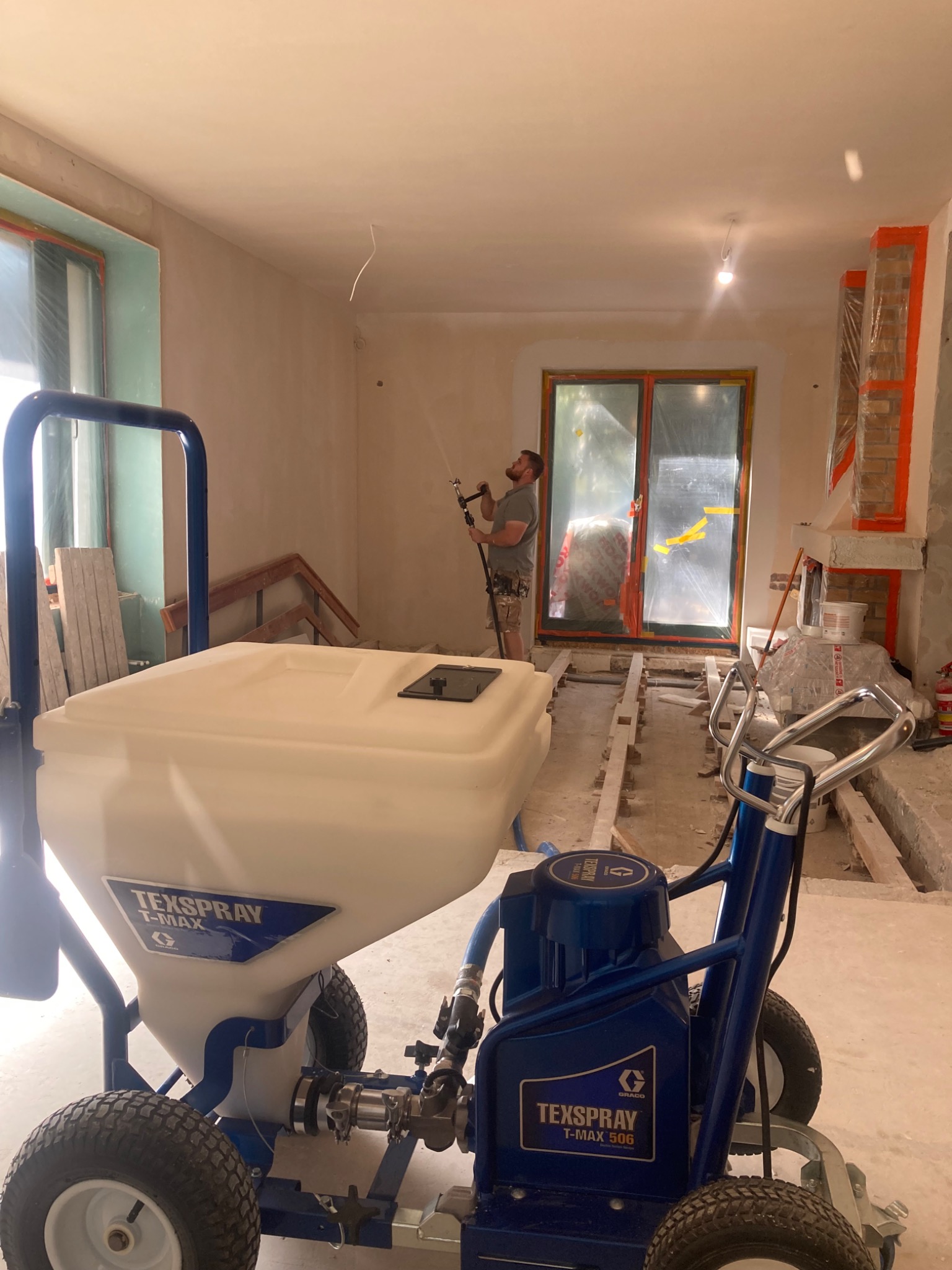 Wnętrze w trakcie prac wykończeniowych, widoczny agregat malarski Texspray T-MAX 506 i pracownik natryskujący ścianę, okno zabezpieczone folią.