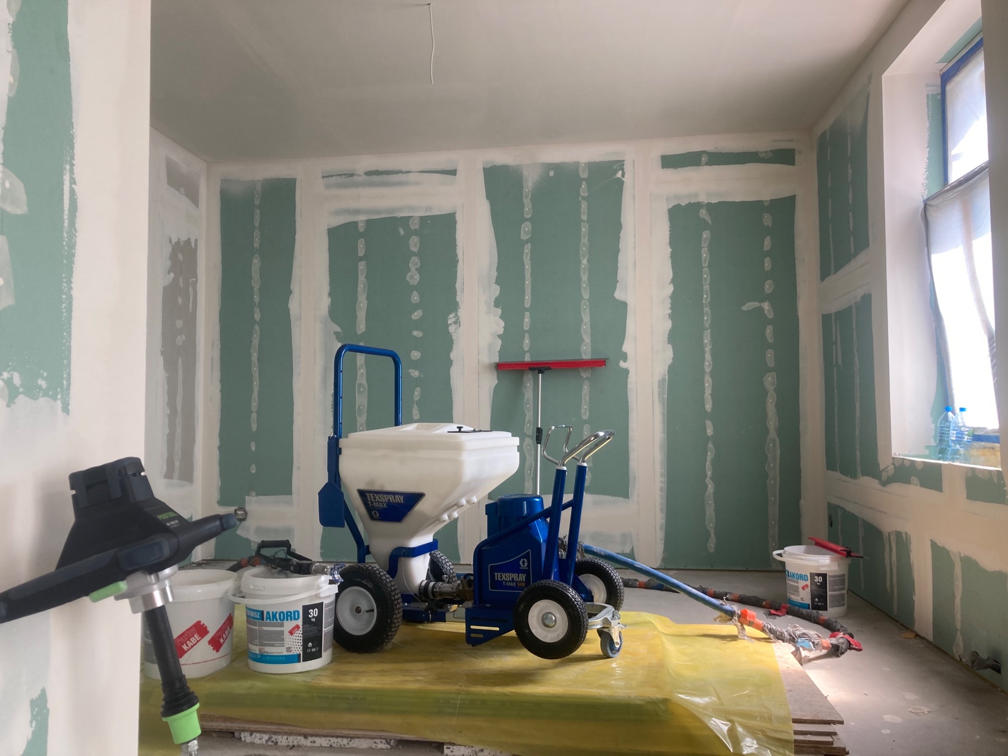 Wnętrze w trakcie prac przygotowawczych do wykończenia ścian, widoczne płyty kartonowo-gipsowe z obrzeżami pokrytymi białą masą, profesjonalny agregat do natrysku masy szpachlowej Texspray, wiadro...