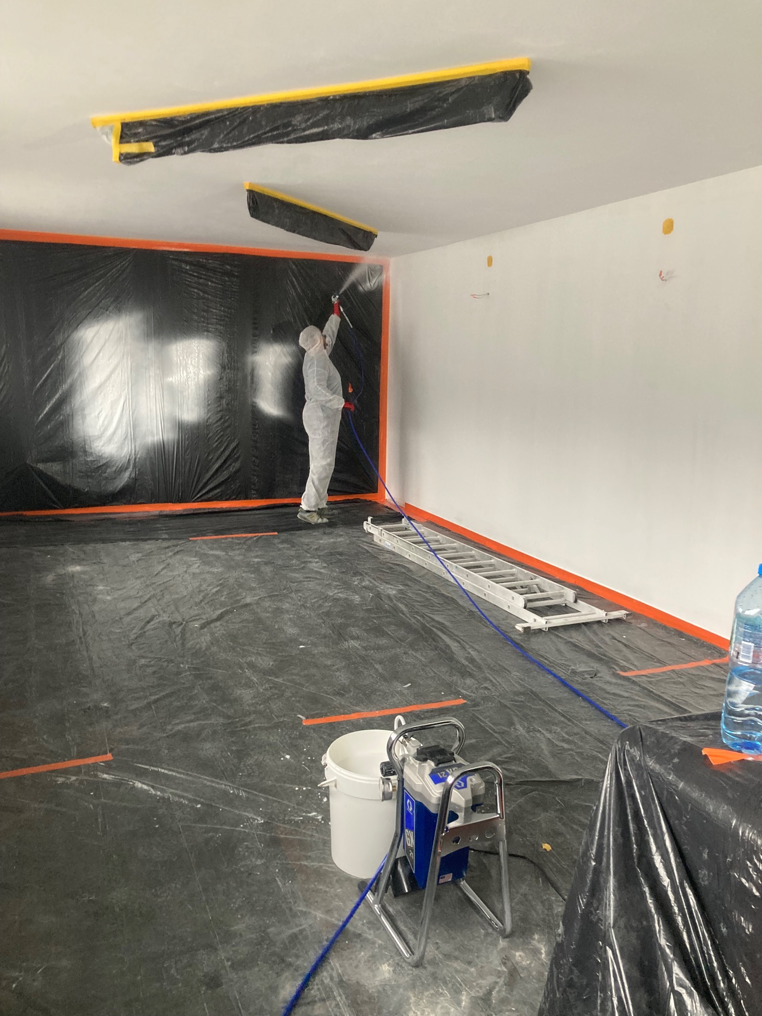 Pracownik w kombinezonie ochronnym maluje ściany agregatem natryskowym w pomieszczeniu zabezpieczonym folią i taśmą malarską, widoczny sprzęt do malowania i drabina.