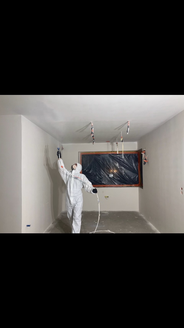 Pracownik w kombinezonie ochronnym maluje ściany w pomieszczeniu za pomocą pistoletu natryskowego, okno zabezpieczone folią.