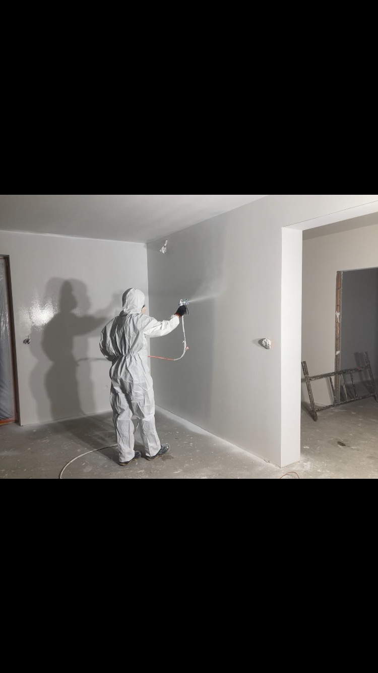 Pracownik w kombinezonie maluje ścianę natryskowo na szaro, widoczny wąż i pistolet natryskowy, w tle surowy stan wykończenia.