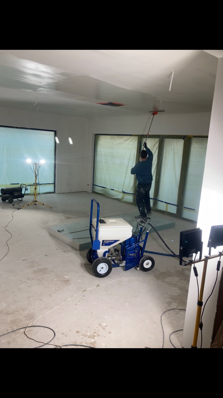 Pracownik w trakcie natryskowego nakładania gładzi na sufit w pomieszczeniu z dużymi oknami zabezpieczonymi folią, widoczny agregat do natrysku i oświetlenie budowlane.