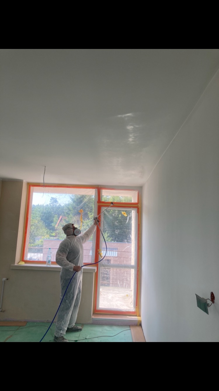 Pracownik w kombinezonie ochronnym maluje natryskowo sufit w pomieszczeniu z oknem zabezpieczonym folią malarską.