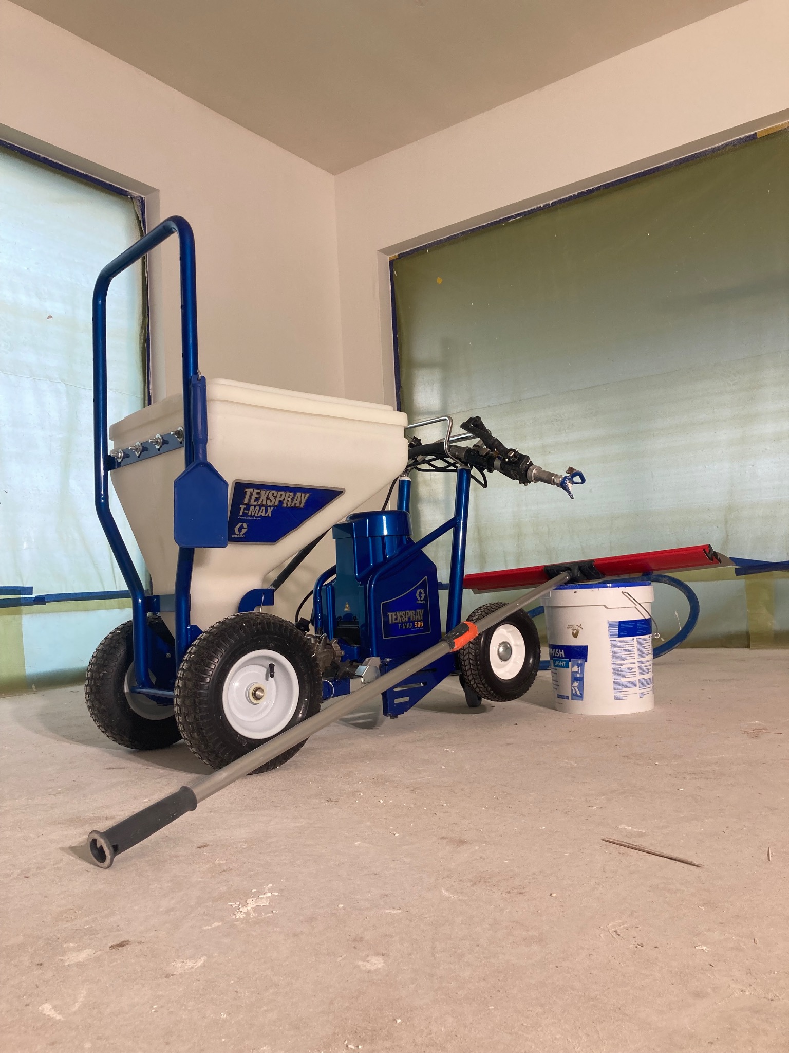 Agregat malarski Texspray T-Max 506 na kółkach z lancą i wiadrem farby w pomieszczeniu z oknami zabezpieczonymi folią.