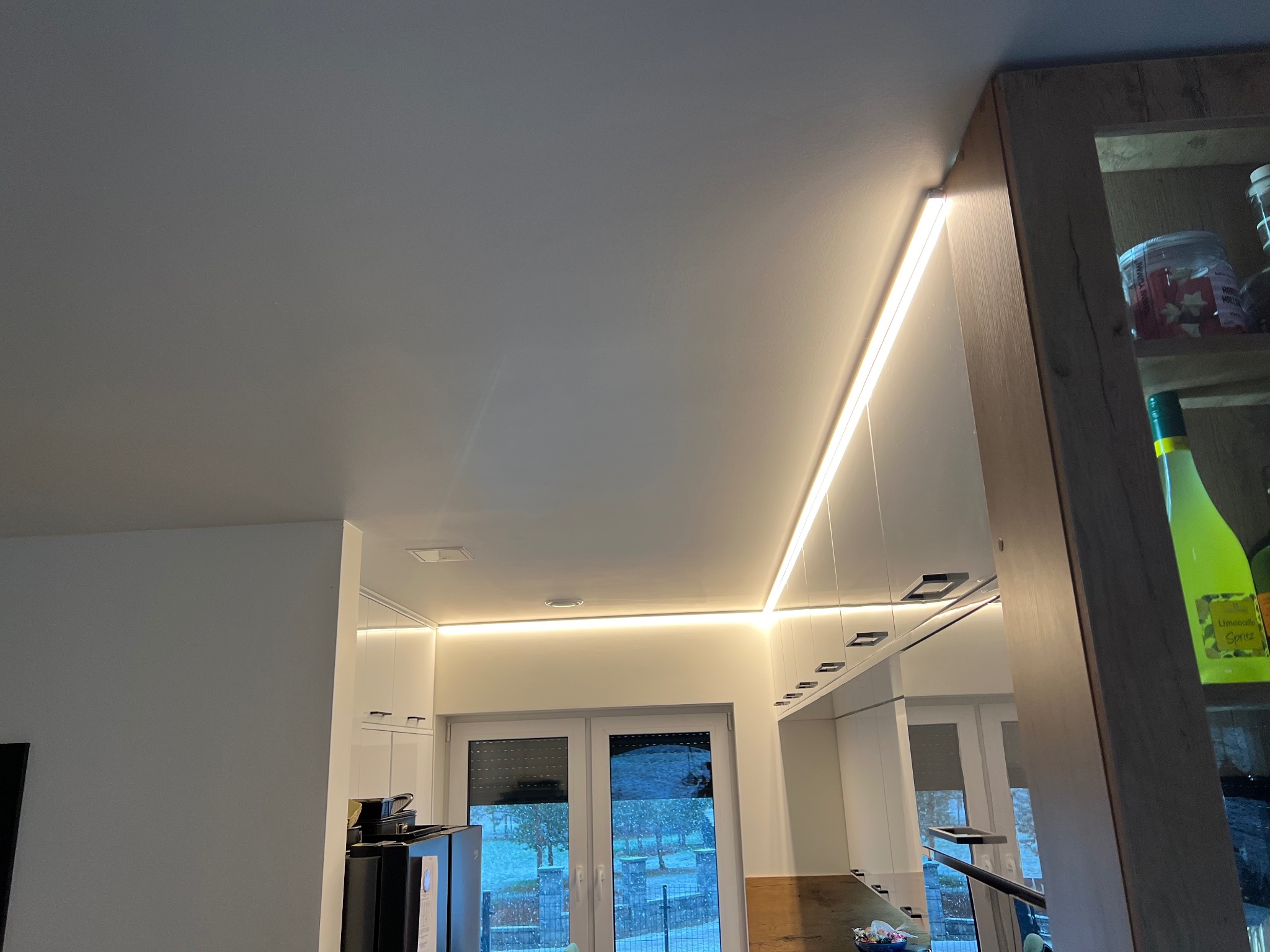 Nowoczesna kuchnia z białymi szafkami i subtelnym oświetleniem LED podszafkowym oraz sufitowym, widok na zewnątrz przez okno w tle.