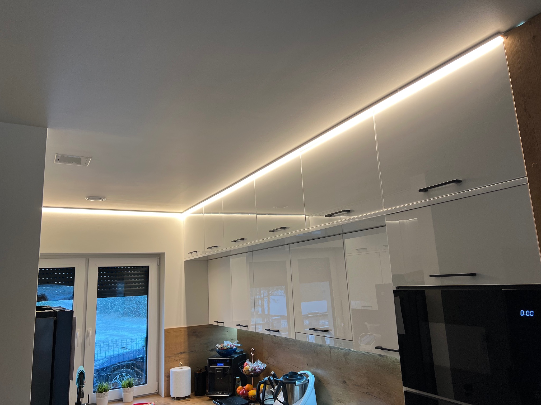 Nowoczesna kuchnia z białymi, błyszczącymi szafkami pod sufitem, oświetlonymi listwą LED, z widocznymi elementami wyposażenia na drewnopodobnym blacie i oknem z roletami w tle.