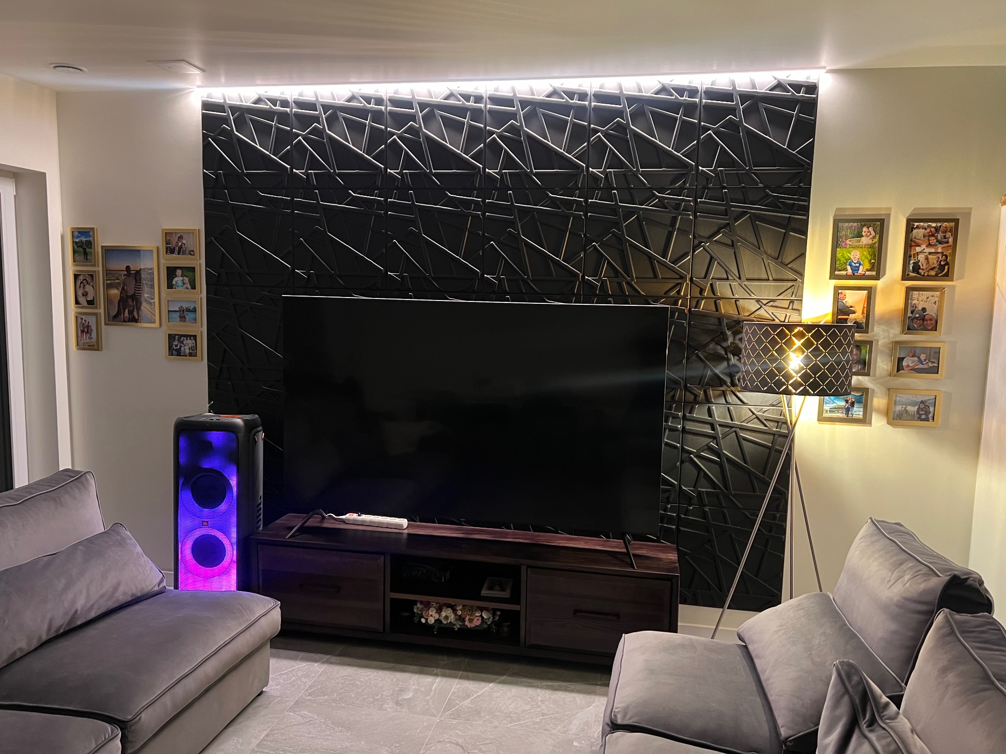 Nowoczesny salon z podświetlaną ścianą 3D, szarym narożnikiem, lampą podłogową i kolumną głośnikową JBL z podświetleniem LED.