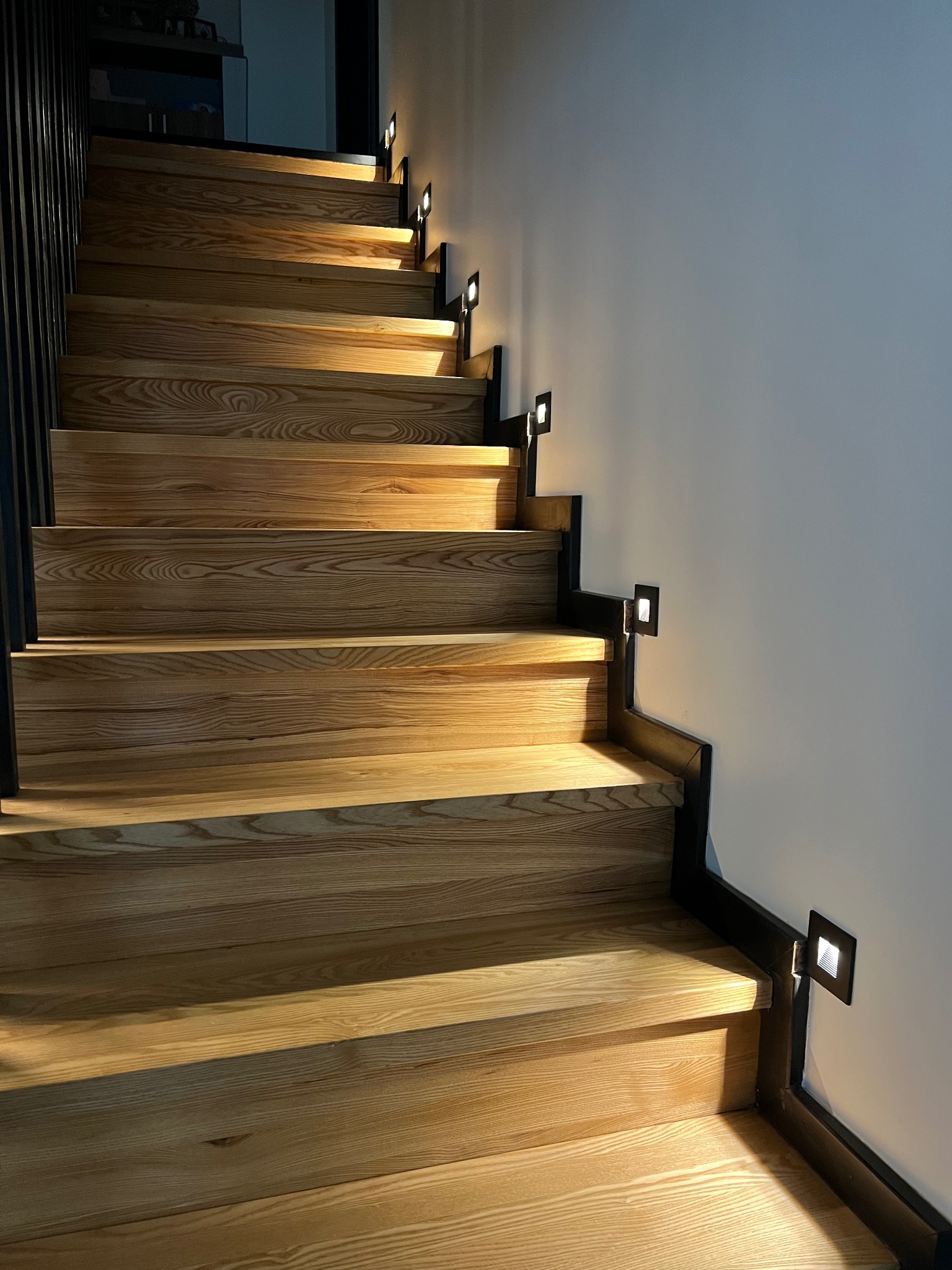 Drewniane schody z zamontowanymi, kwadratowymi lampkami LED w czarnych obudowach, oświetlającymi stopnie. Lampki umieszczone są wzdłuż boku schodów, na ścianie.
