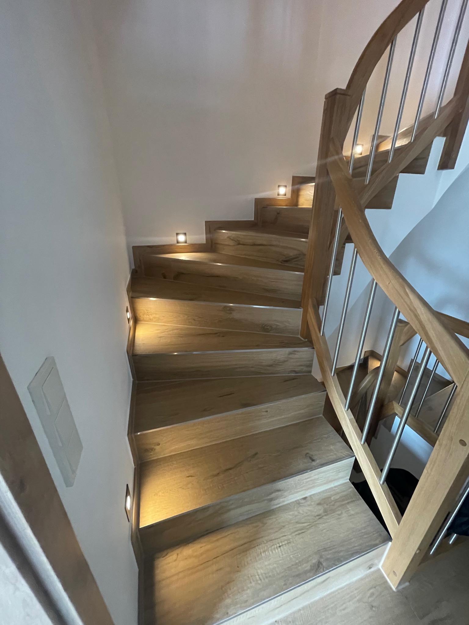 Drewniane schody z zamontowanymi punktowymi lampkami LED w ścianie i stopniach, oświetlające bieg schodów. Balustrada wykonana z drewna i metalowych prętów.