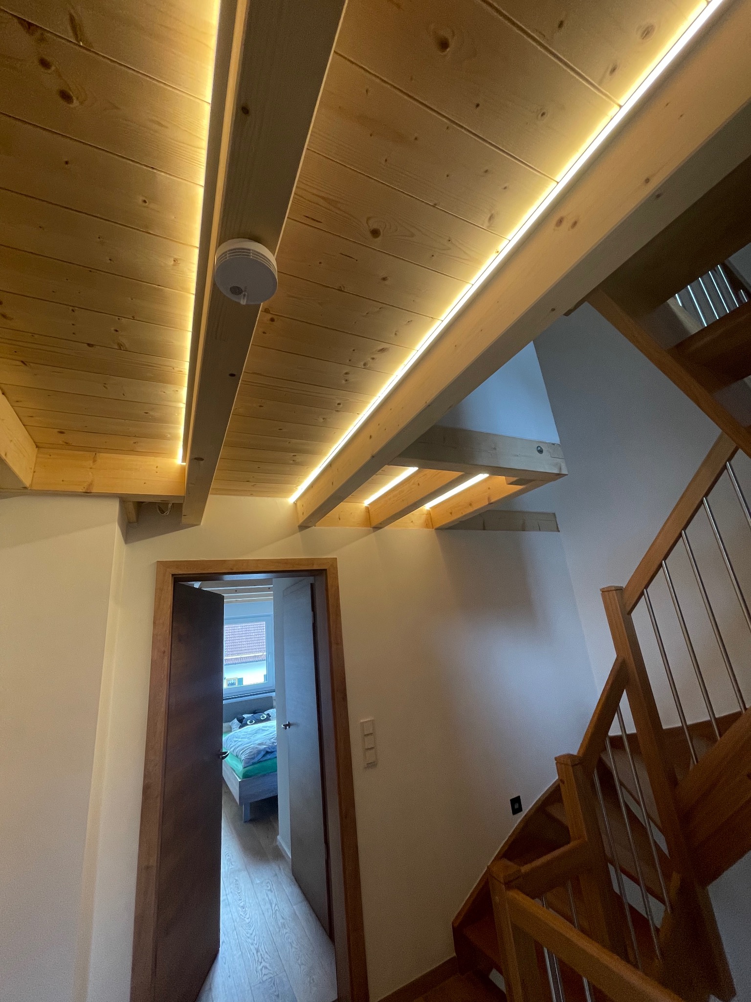 Drewniany sufit poddasza z zamontowanym oświetleniem LED liniowym, widoczny czujnik dymu, fragment schodów z drewnianą balustradą i stalowymi prętami oraz otwarte drzwi do sypialni.