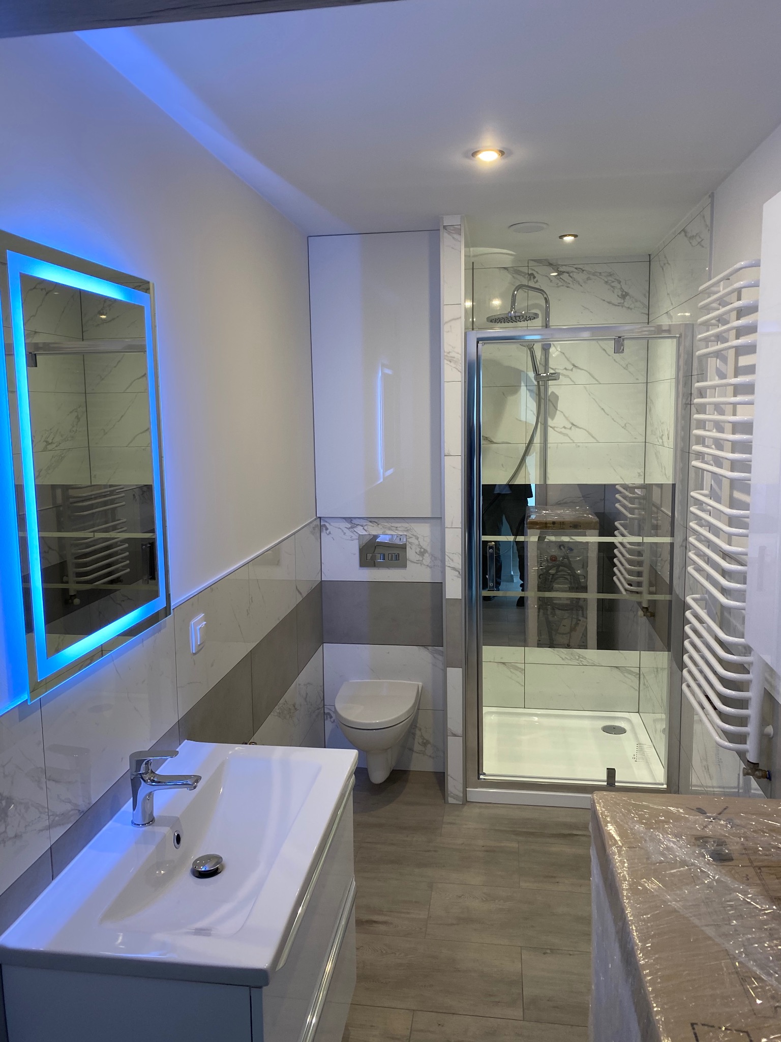 Nowoczesna łazienka z podświetlanym lustrem LED, kabiną prysznicową z chromowanymi detalami i grzejnikiem drabinkowym, wykończona płytkami imitującymi marmur