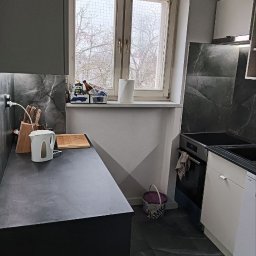 Przemysław Stefański - Częściowy widok wykończonej kuchni z czarnym blatem, marmurowymi płytkami na ścianie, oknem z widokiem na drzewa i wiadrem z farbą na podłodze.