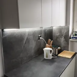 Nowoczesna kuchnia z szarym blatem i marmurową ścianą, oświetlenie LED pod szafkami, biały czajnik elektryczny i drewniana deska do krojenia na blacie.