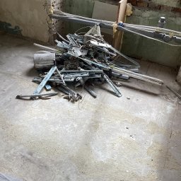 Przemysław Stefański - Stare elementy metalowe i rury leżące na betonowej podłodze w trakcie demontażu w pomieszczeniu z oknem i widocznymi cegłami.