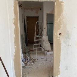 Przemysław Stefański - Przygotowanie otworu drzwiowego do osadzenia ościeżnicy, widoczne obrobione krawędzie ścian i surowy stan wykończenia wnętrza z widoczną drabinką i resztkami materiałów budowlanych.