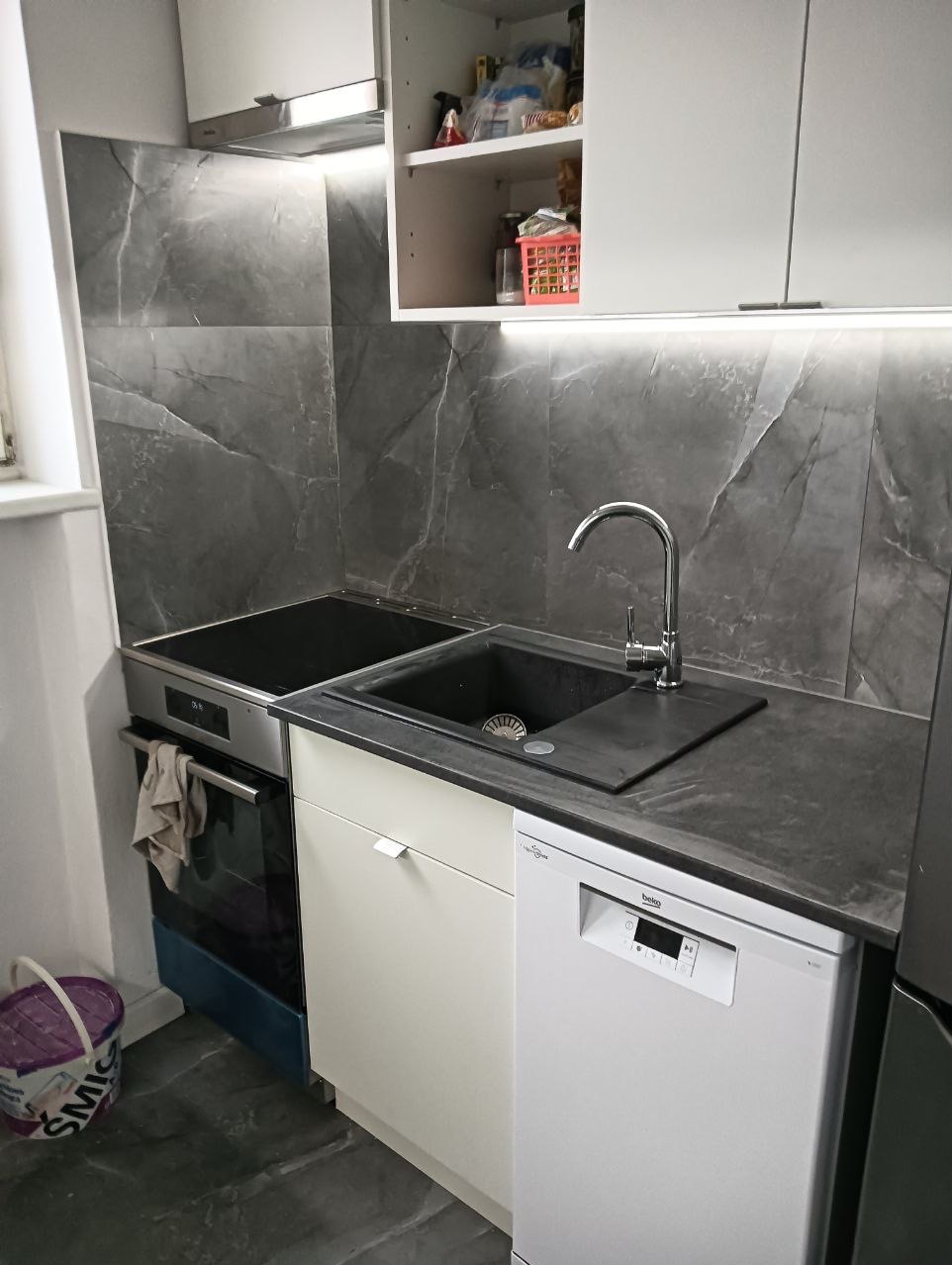 Nowoczesna kuchnia z szarymi płytkami imitującymi marmur na ścianie, czarnym zlewem i blatem, białą zabudową szafek, zmywarką Beko i płytą indukcyjną, oświetlona ledowym pasem pod szafkami wiszącymi.