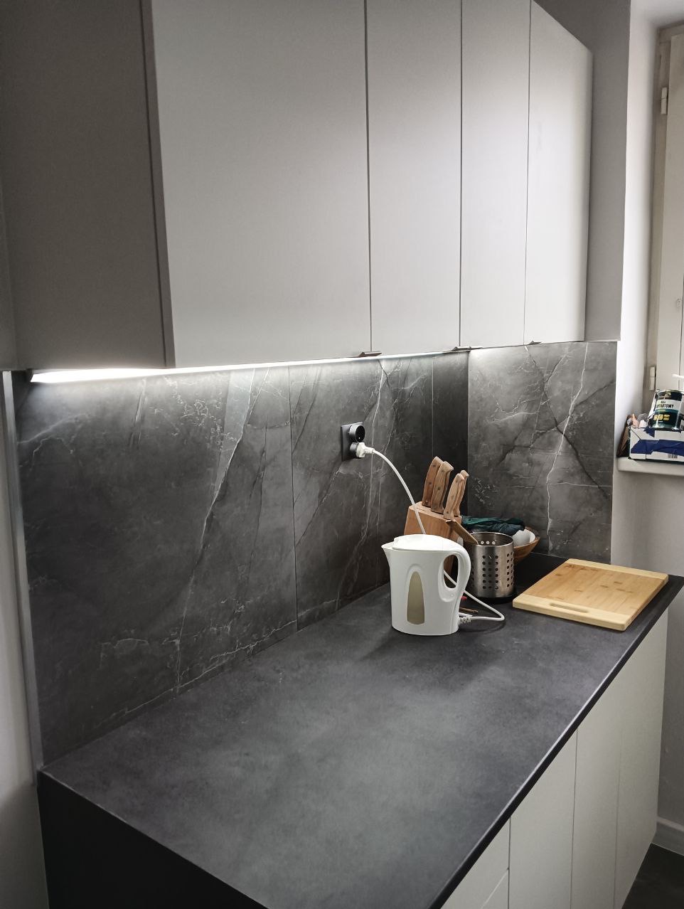 Nowoczesna kuchnia z szarym blatem i marmurową ścianą, oświetlenie LED pod szafkami, biały czajnik elektryczny i drewniana deska do krojenia na blacie.