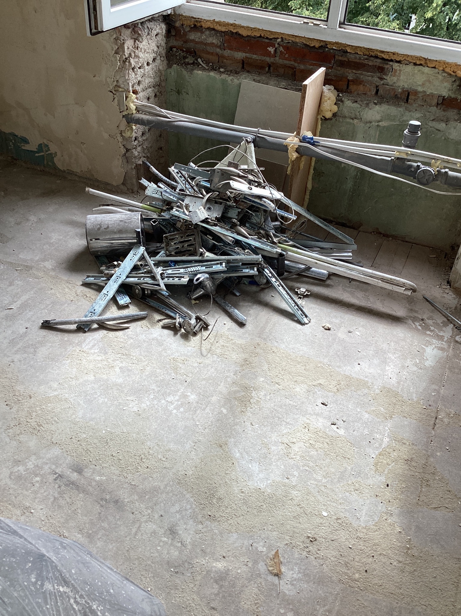 Stare elementy metalowe i rury leżące na betonowej podłodze w trakcie demontażu w pomieszczeniu z oknem i widocznymi cegłami.