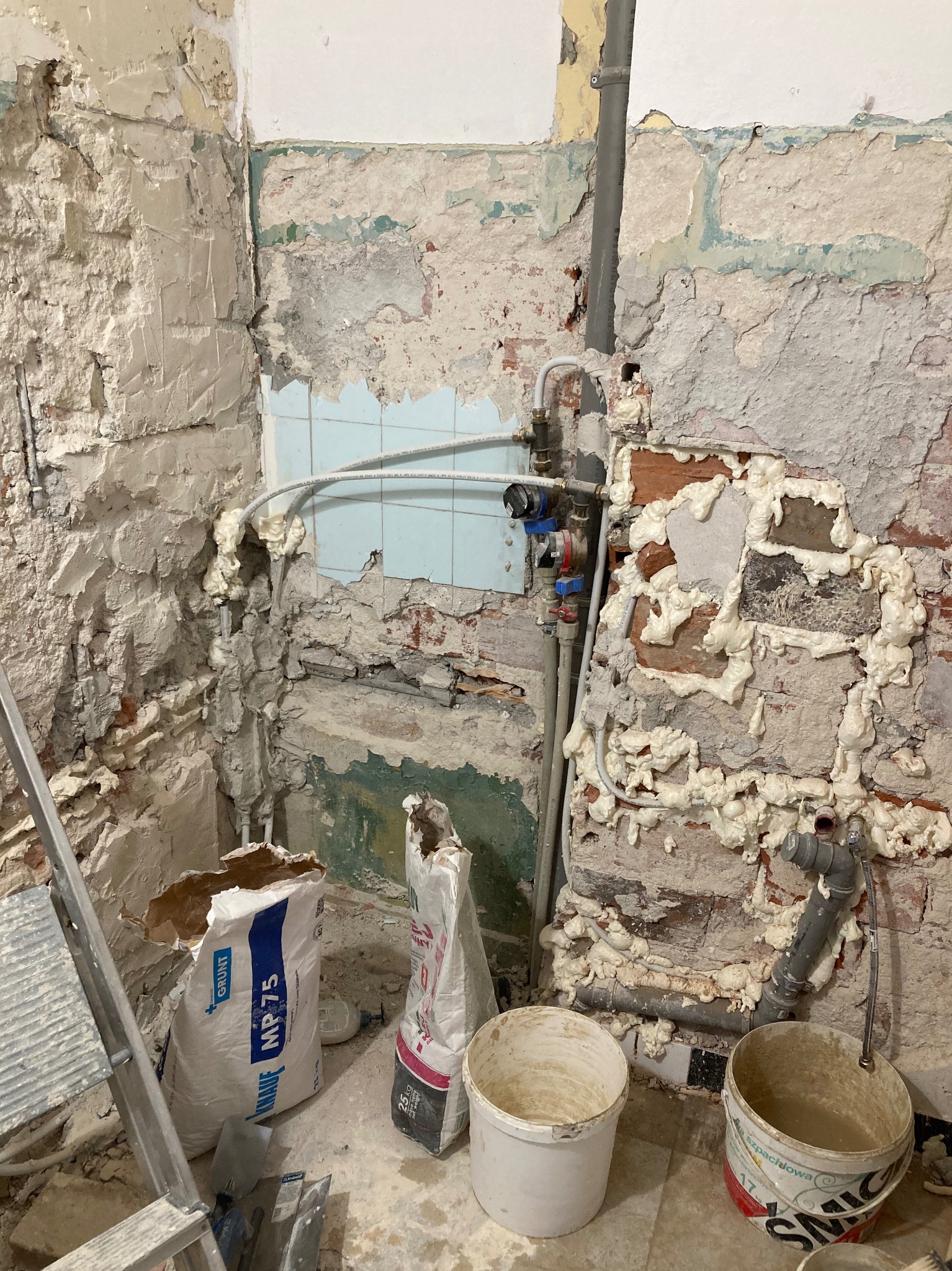 Fragment ściany w trakcie prac remontowych, widoczne odsłonięte cegły, instalacje, piana montażowa, worki z gruntem i zaprawą oraz drabina malarska.