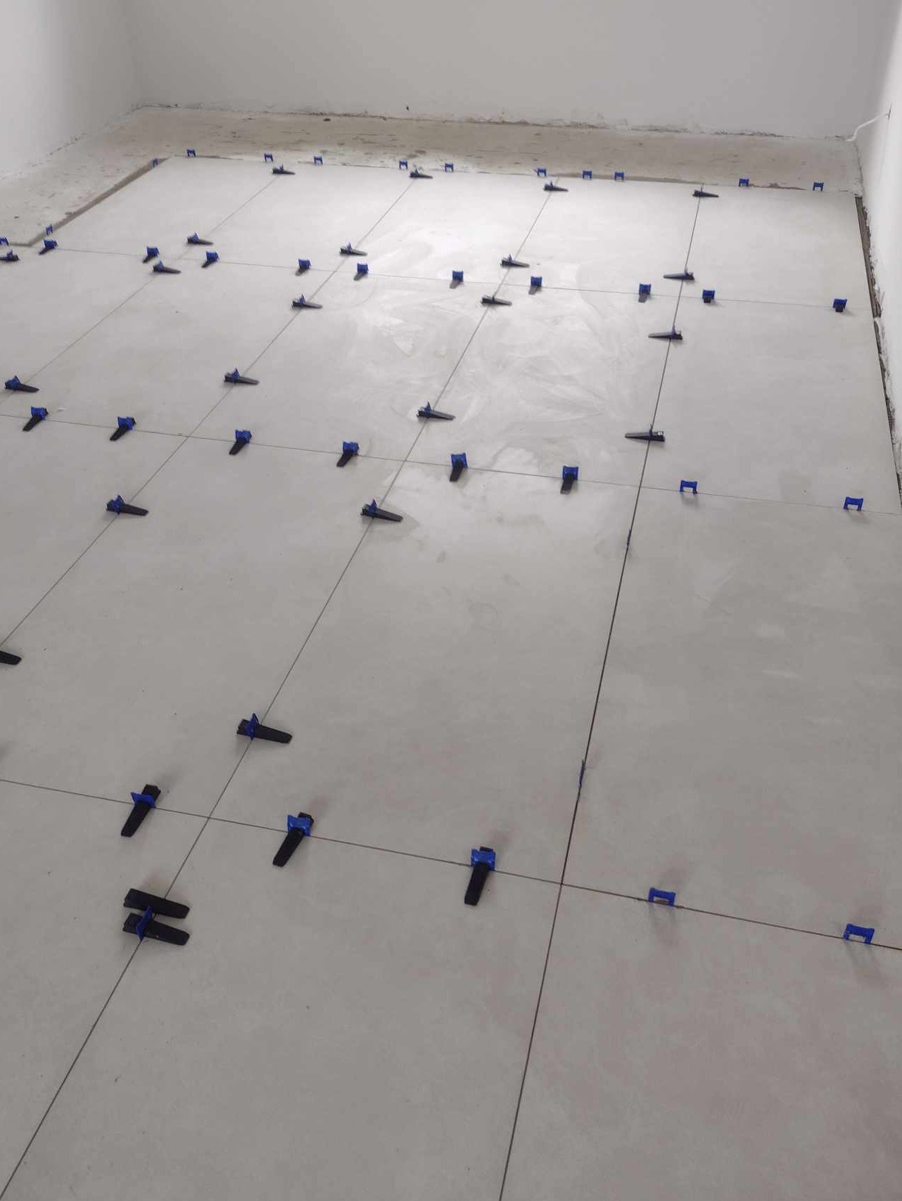 Świeżo ułożone, jasne płytki podłogowe z niebieskimi klipsami poziomującymi i sznurkami wyznaczającymi linie fug, w trakcie prac wykończeniowych.