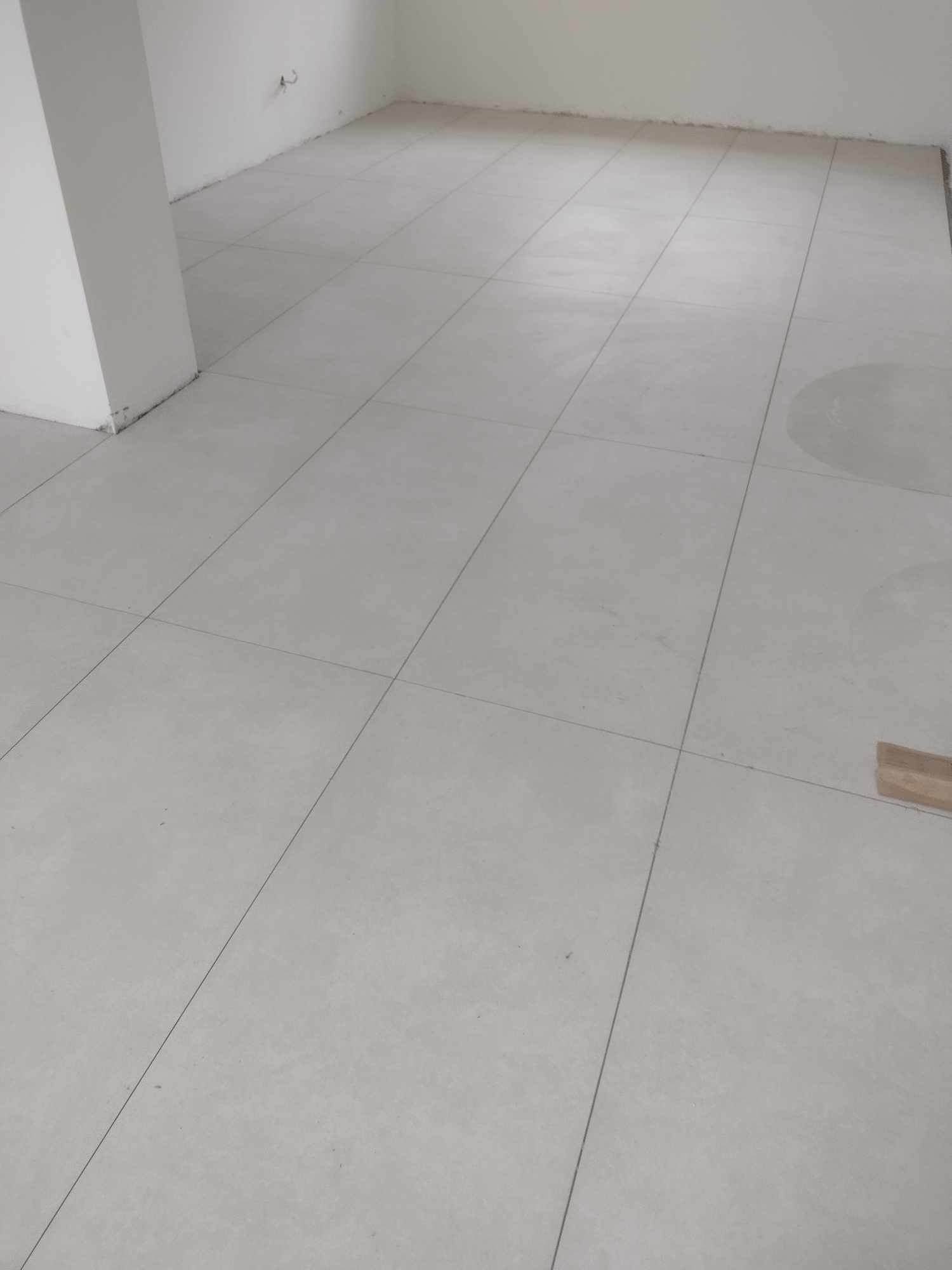 Jasnoszara, matowa podłoga wyłożona płytkami w pustym pomieszczeniu, widoczny fragment listwy przypodłogowej i ślady po ołówku na fugach.