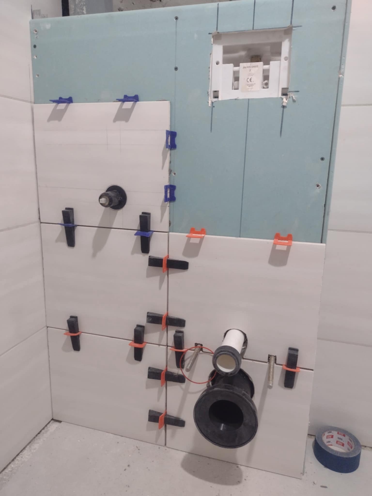 Układanie białych płytek na ścianie z widocznymi elementami instalacji hydraulicznej i poziomującymi klipsami, fragment płyty gipsowej z wycięciem na instalację, niebieska taśma malarska w rogu