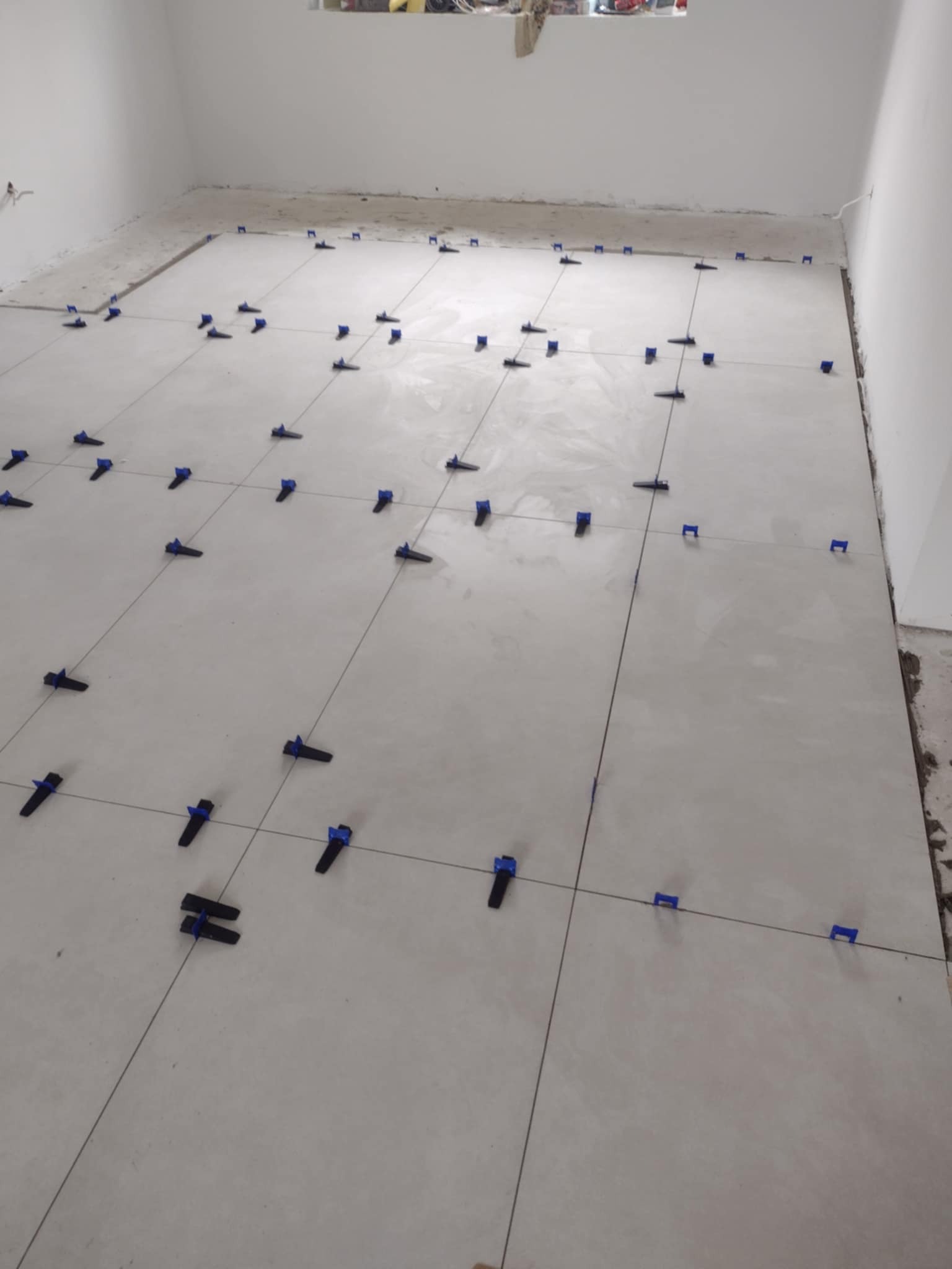 Układanie jasnoszarych płytek podłogowych w pustym pomieszczeniu z użyciem czarnych i niebieskich klipsów poziomujących, widoczne fugi i surowy brzeg podłogi przy ścianie.