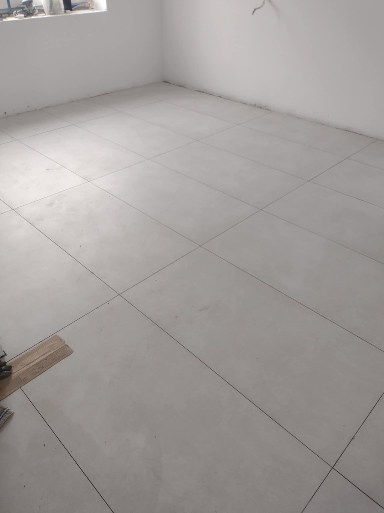 Świeżo ułożone, jasnoszare płytki podłogowe z widocznymi fugami w pustym pomieszczeniu, z narzędziami do układania płytek w lewym dolnym rogu.