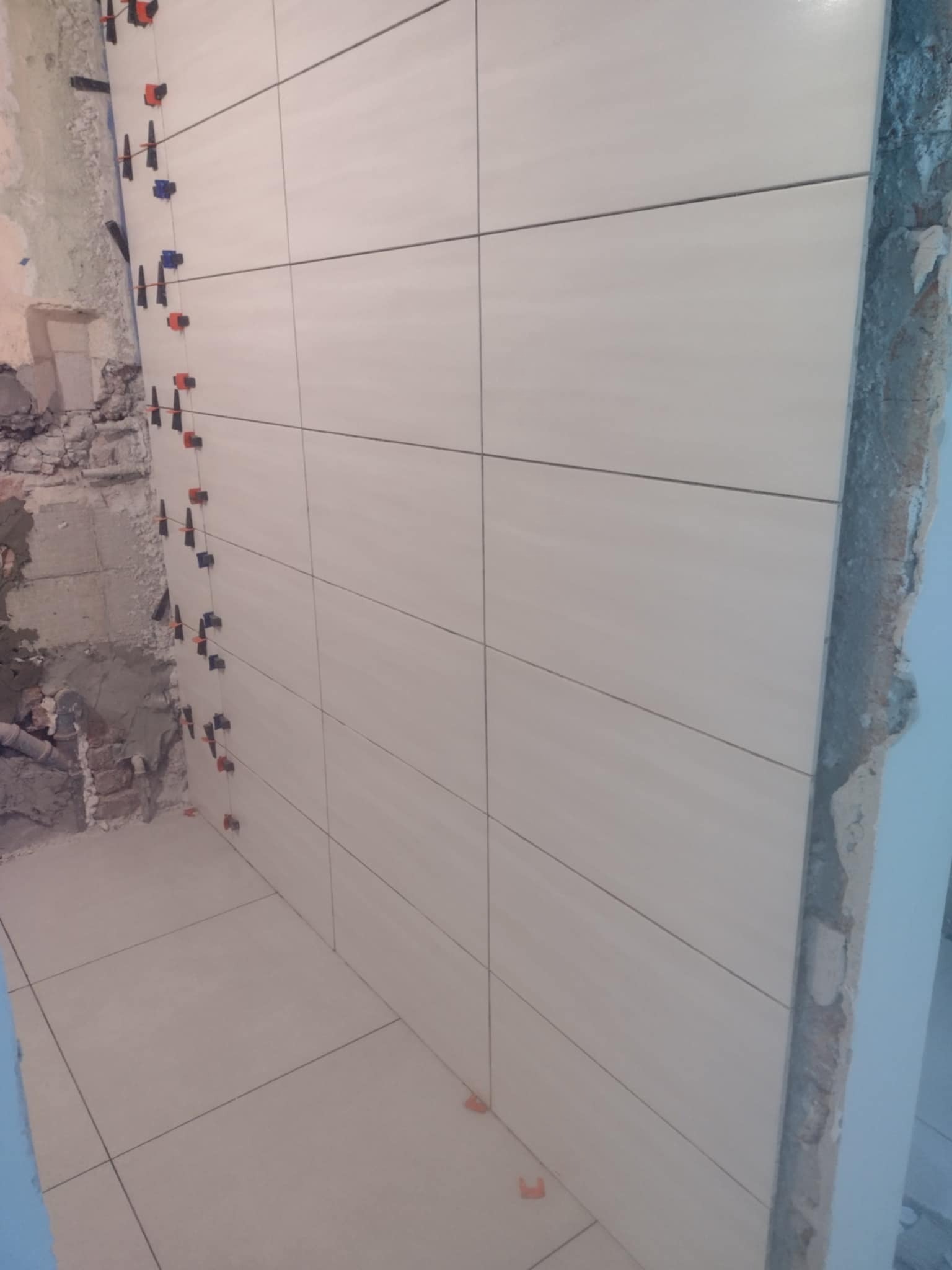 Ściana i podłoga w trakcie układania jasnych, prostokątnych płytek z użyciem klipsów poziomujących; widoczne surowe fragmenty ściany.