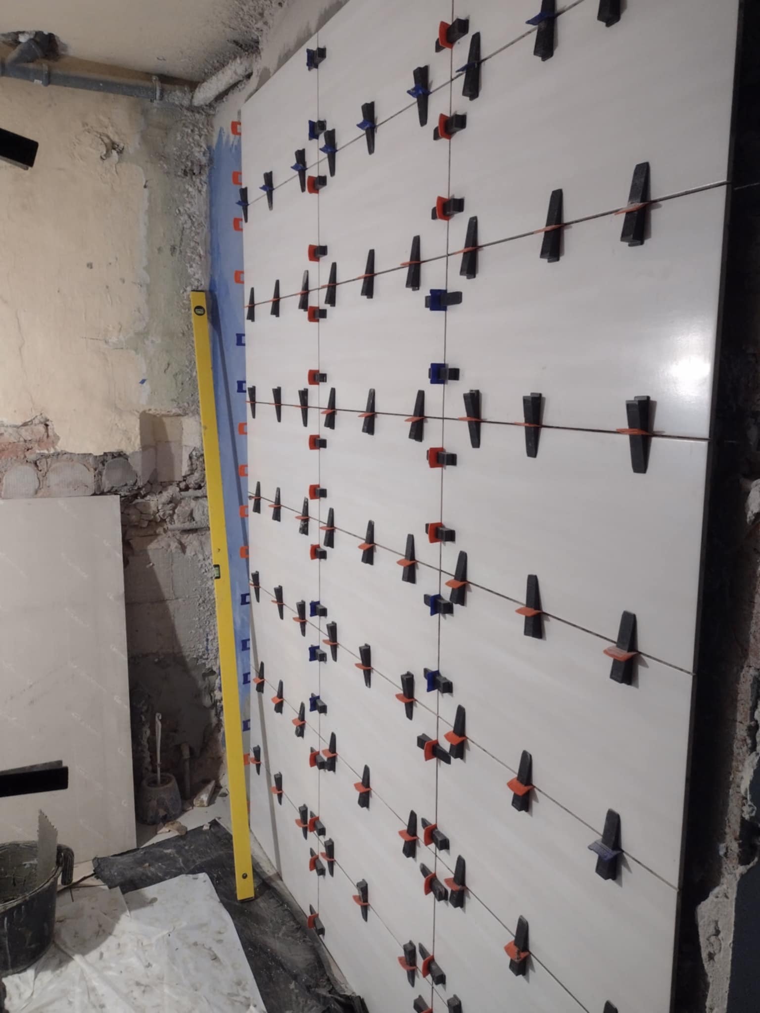 Układanie białych płytek na ścianie w Radomsku z użyciem systemu poziomowania płytek (klipsy i kliny), widoczna poziomica i przygotowane podłoże.