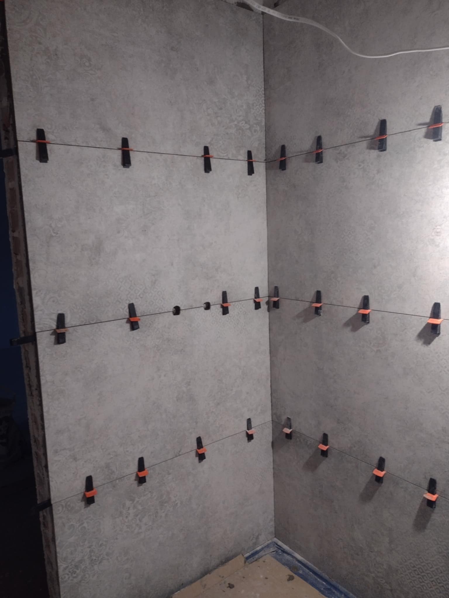 Świeżo położone szare płytki ścienne w rogu pomieszczenia, wyrównane za pomocą czarnych klipsów z pomarańczowymi wstawkami i żyłki murarskiej, widoczny fragment podłogi.