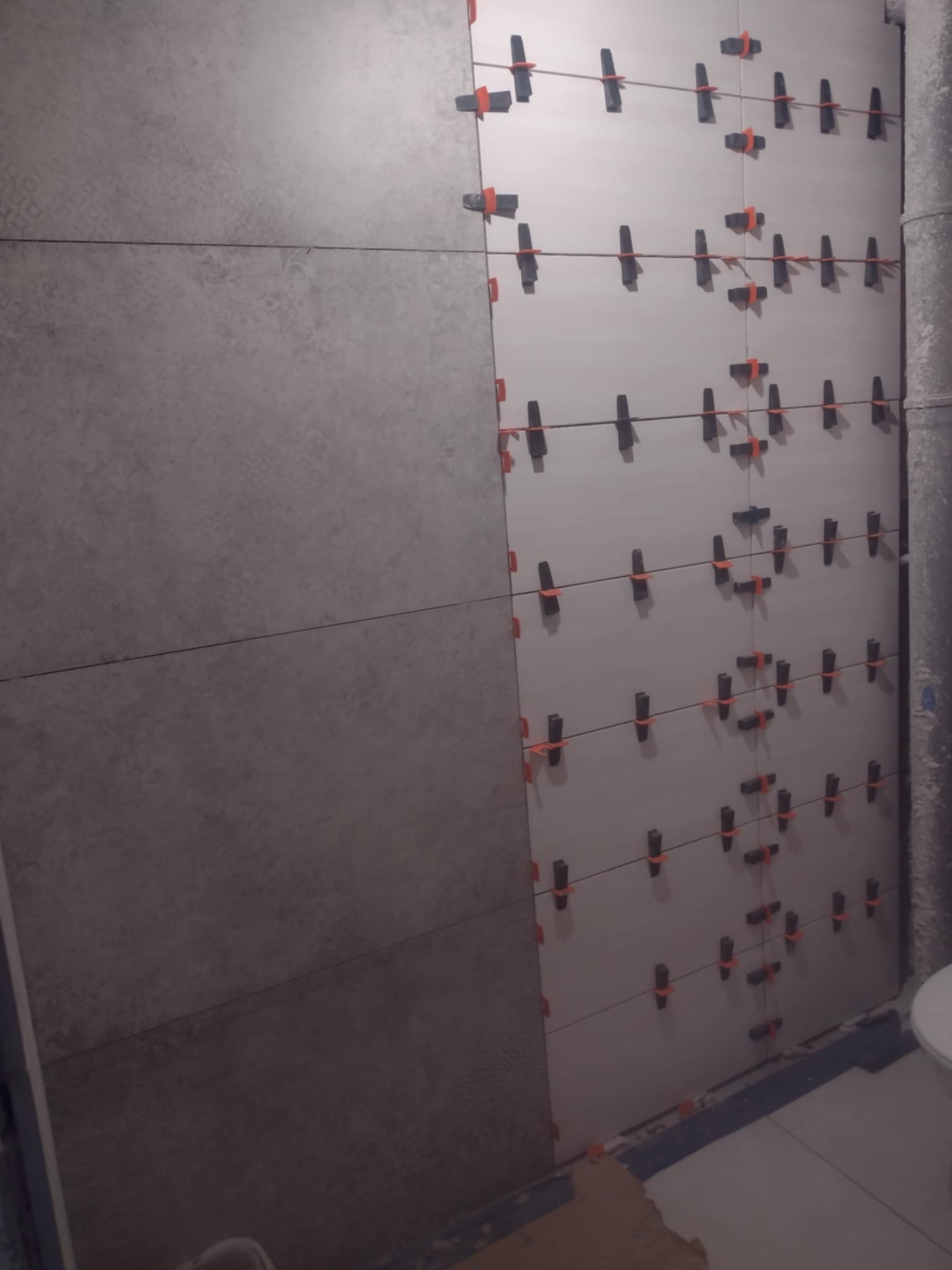 Ściana w trakcie układania płytek z użyciem klipsów poziomujących, widoczny fragment toalety oraz rura kanalizacyjna.