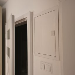 MLT IT Paweł Góral - Biała skrzynka rozdzielcza z bezpiecznikami na ścianie w mieszkaniu w Warszawie, obok włączniki światła i częściowo otwarte drzwi z szybkami.