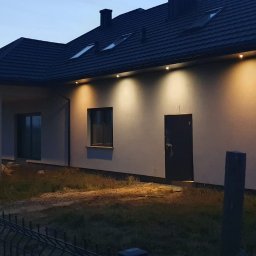 MLT IT Paweł Góral - Nowoczesny dom jednorodzinny z oświetleniem zewnętrznym LED, widoczny wieczorem, z czarną dachówką i grafitową stolarką okienną, ogrodzony metalowym płotem.