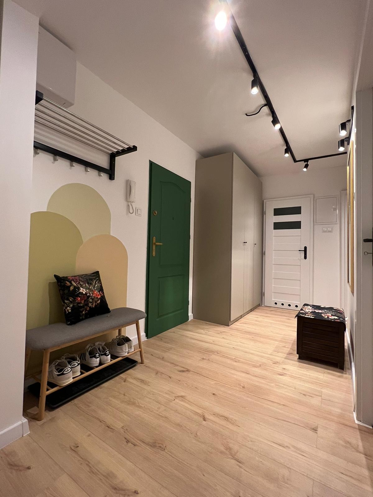 Nowoczesny przedpokój z drewnianą podłogą, zielonymi drzwiami, szarą szafą, oświetleniem punktowym na suficie, wieszakiem na ścianie, ławką z poduszką i półką na buty.