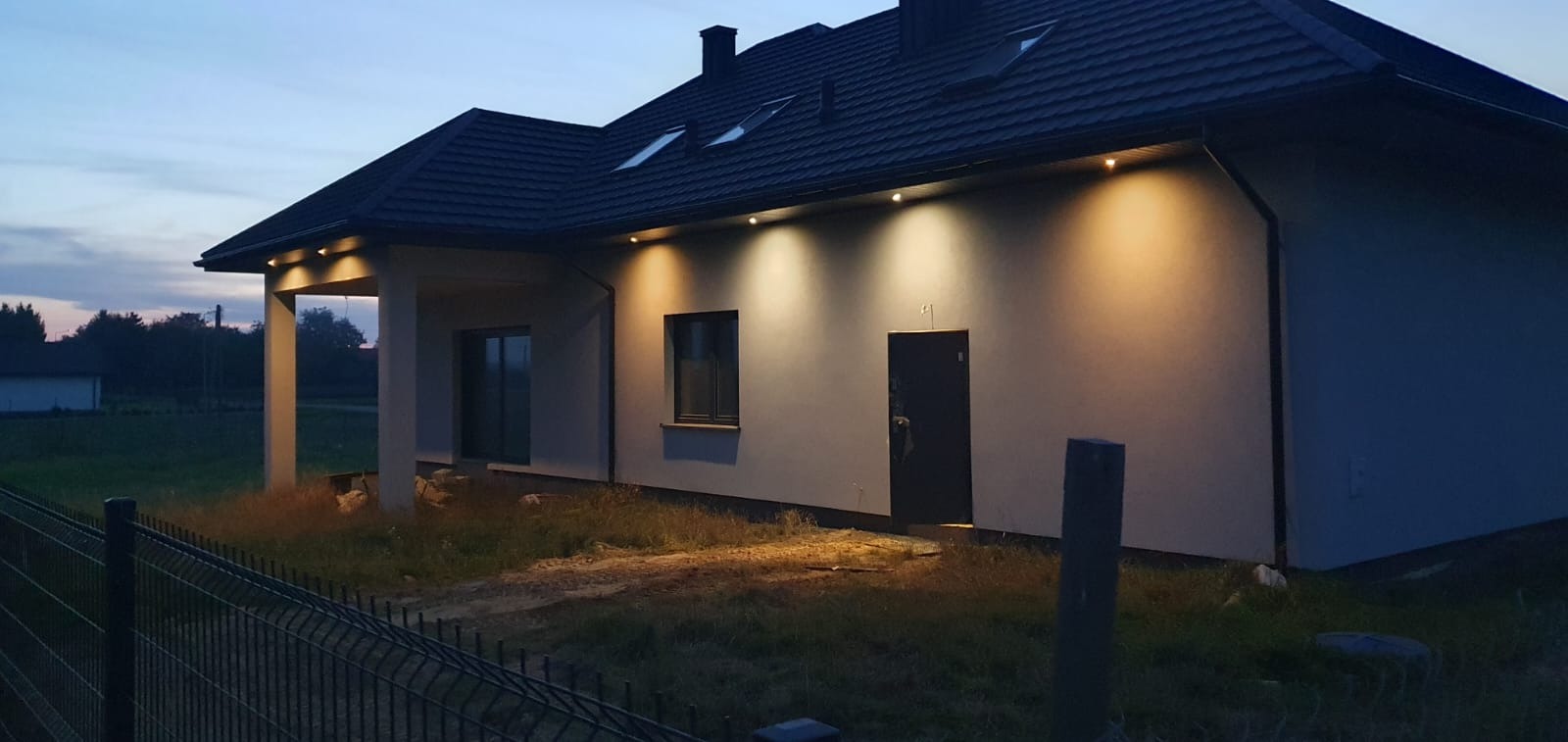 Nowoczesny dom jednorodzinny z oświetleniem zewnętrznym LED, widoczny wieczorem, z czarną dachówką i grafitową stolarką okienną, ogrodzony metalowym płotem.