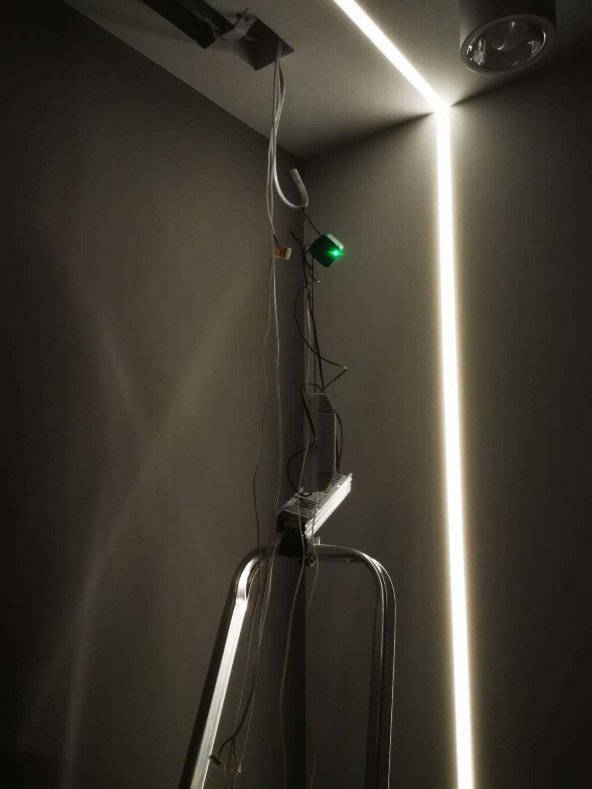 Montaż liniowego oświetlenia LED w rogu pomieszczenia, widoczna drabina, przewody i sterownik z zieloną diodą w trakcie instalacji.